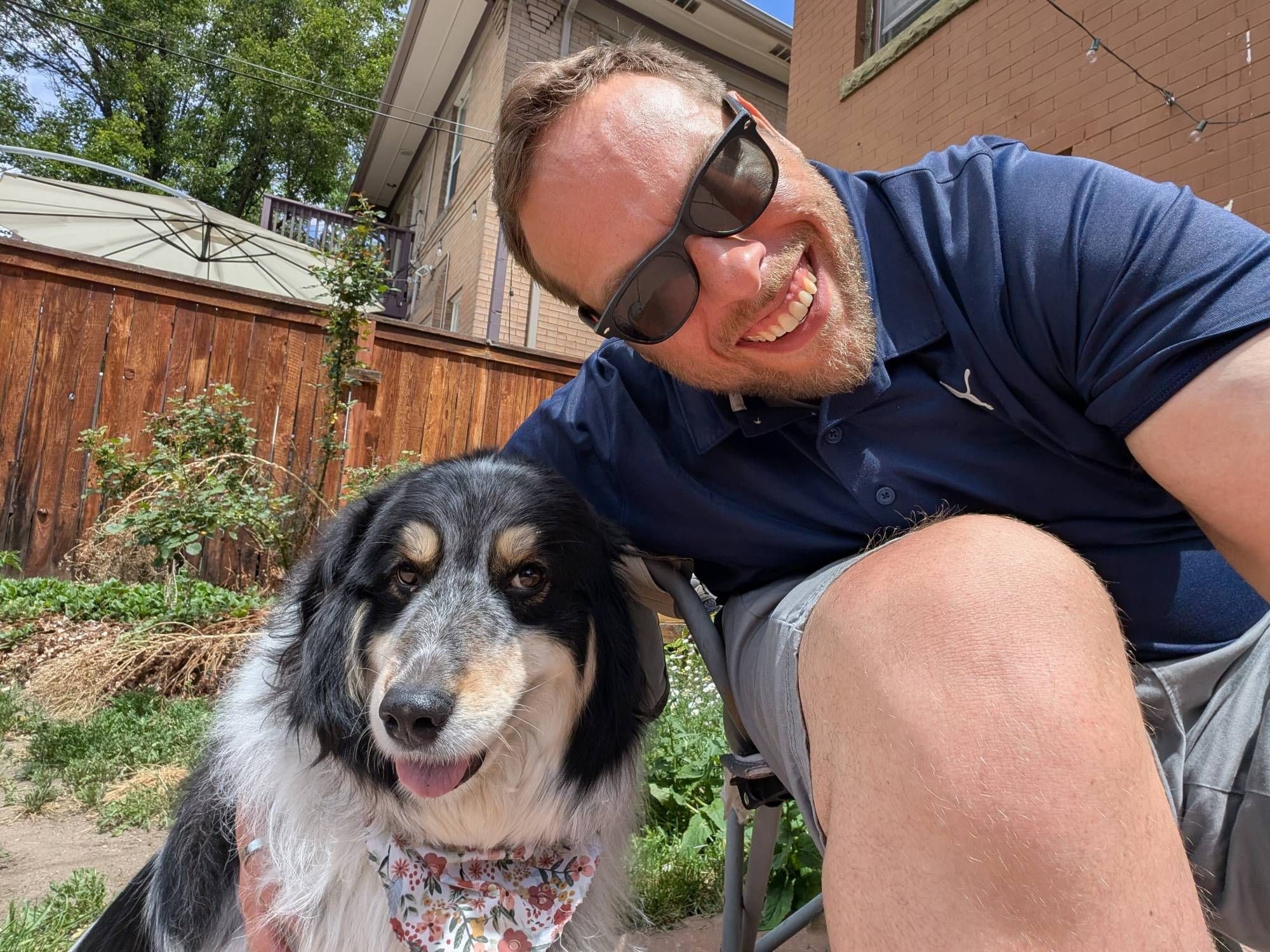 Mike in sunglasses, smiling next to his dog Penny, who wears a floral bandana, in a sunny backyard